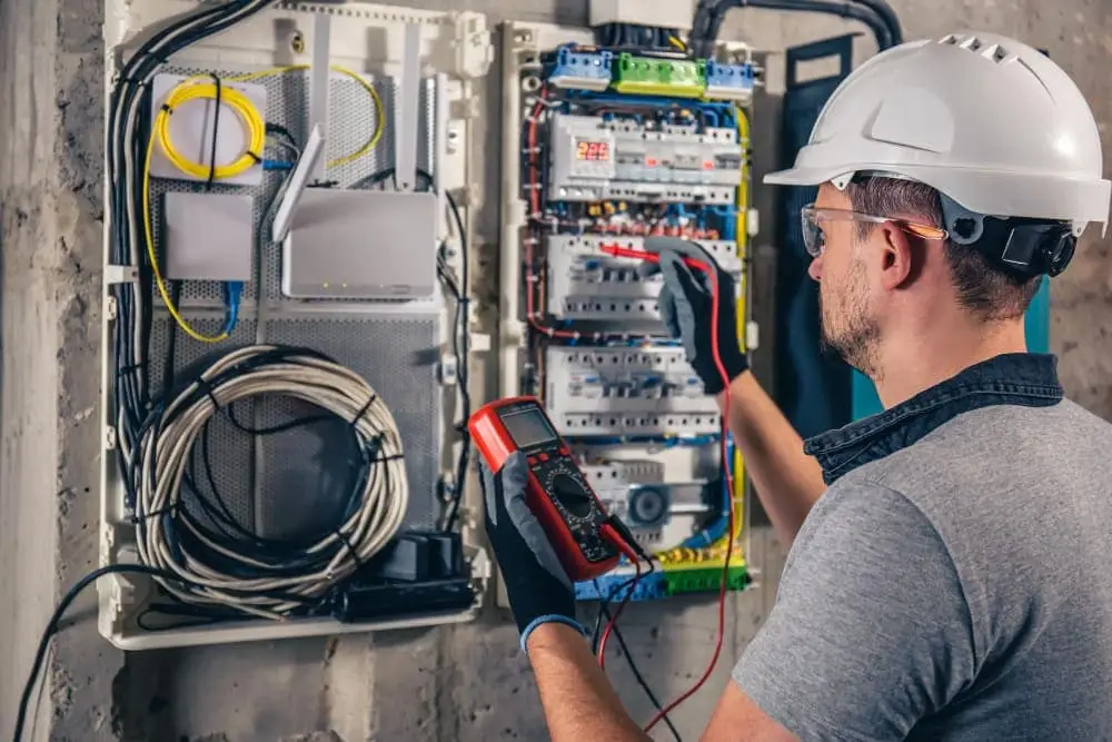 En elektriker med skyddshjälm och glasögon testar en elpanel med en multimeter.