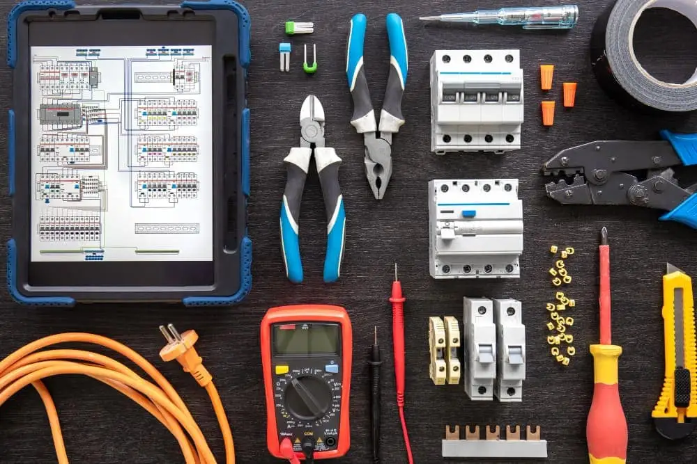 En samling elektriker-verktyg på en mörk yta, inklusive en surfplatta med elscheman, multimeter, skruvmejslar, tänger, brytare, kablar och eltejp.