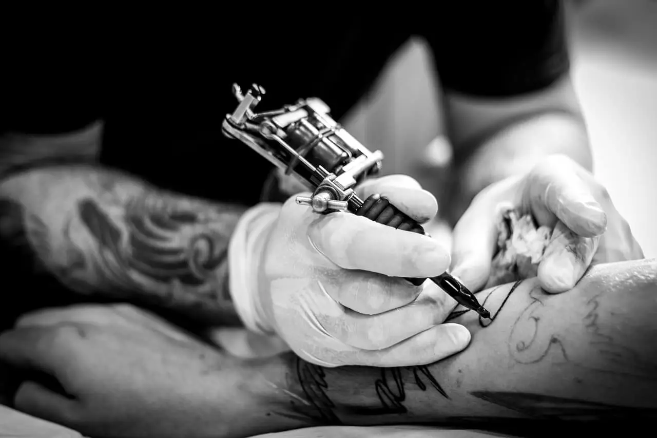 En svartvit bild visar en tatuerare med handskar som tatuerar en persons arm.