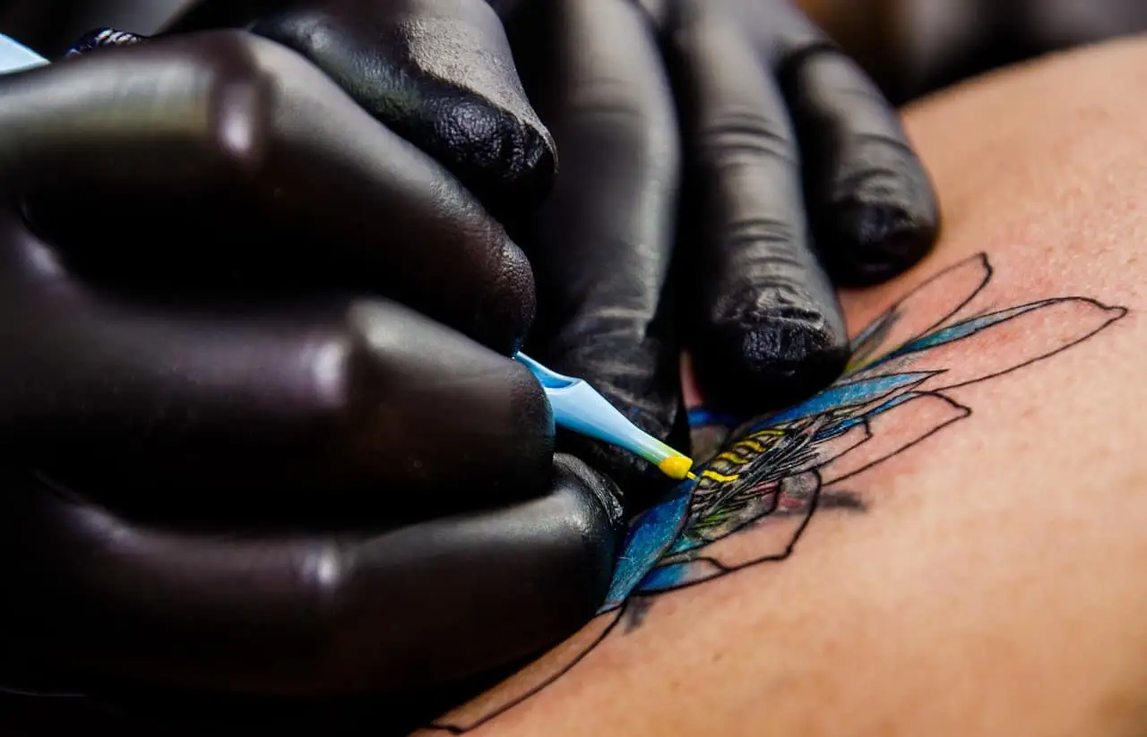 En närbild visar en person med svarta handskar som tatuerar en fjärilsdesign.